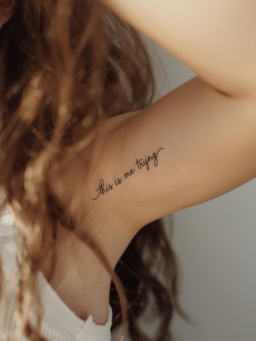 A close-up of a person’s upper arm with a tattoo in cursive script that reads “this is me trying.” Curly hair partially frames the arm, and the person is wearing a white sleeveless top.