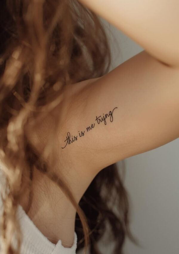 A close-up of a person’s upper arm with the handwritten-style tattoo “this is me trying.” The person has wavy brown hair and is wearing a white sleeveless top.