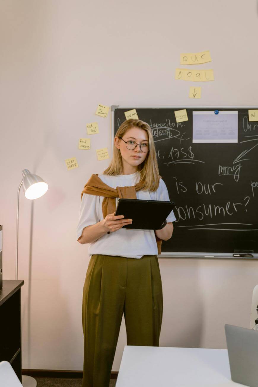 A woman with glasses stands in front of a chalkboard covered in writing and sticky notes, holding a tablet. She wears a white shirt, brown sweater over her shoulders, and olive green pants in a classroom or office setting.