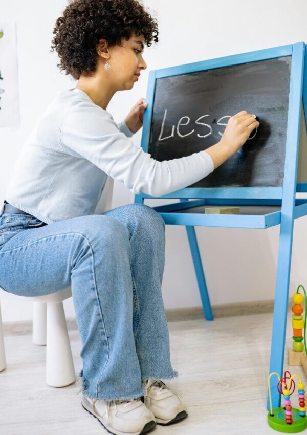 A person with curly hair and jeans writes “LESS” on a small black chalkboard, seated on a white stool. Educational toys and alphabet posters are visible in the background.