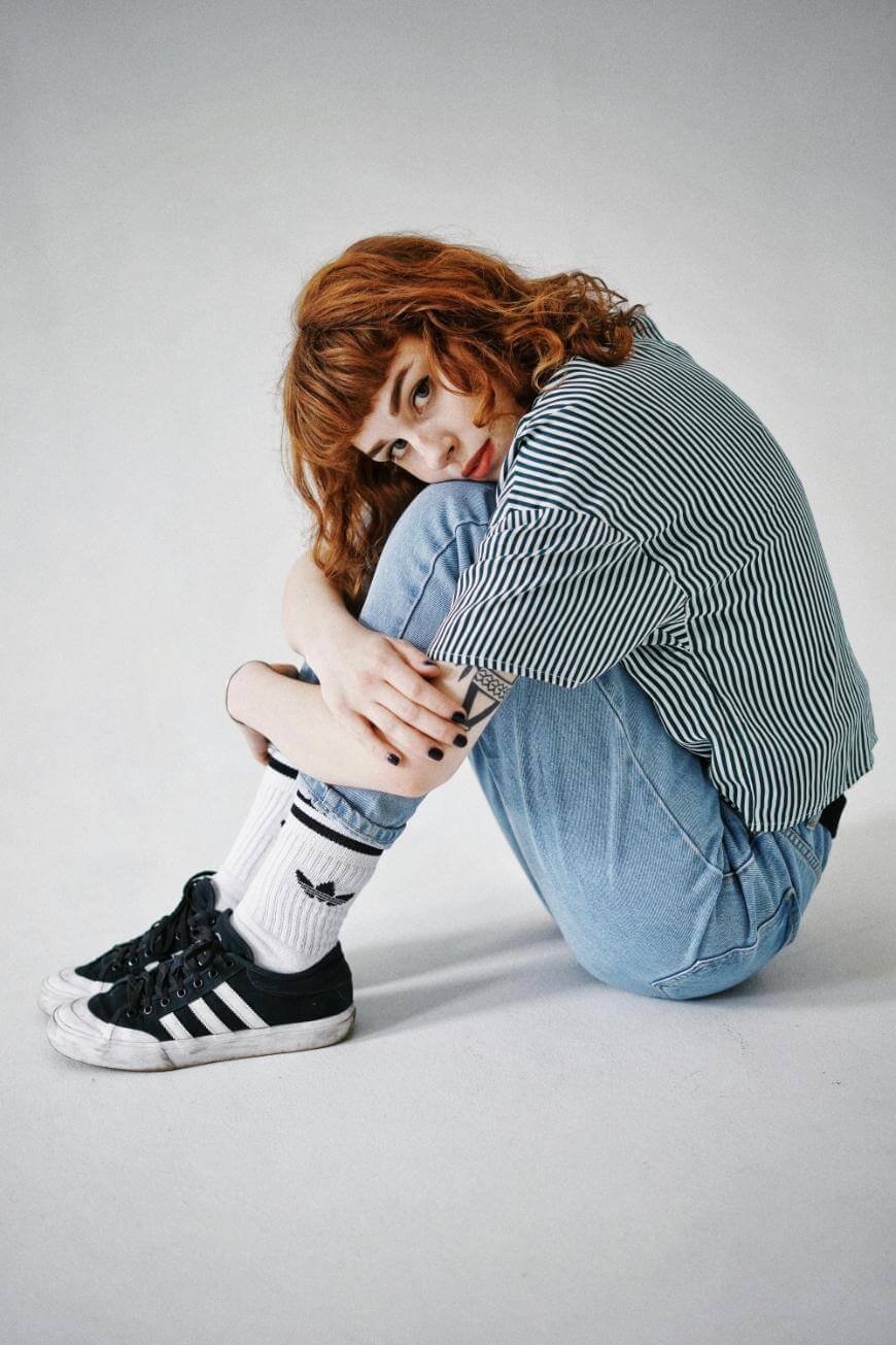 A person with curly red hair sits on the floor hugging their knees, wearing a black-and-white striped shirt, light blue jeans, white socks with designs, and black Adidas sneakers, looking toward the camera.