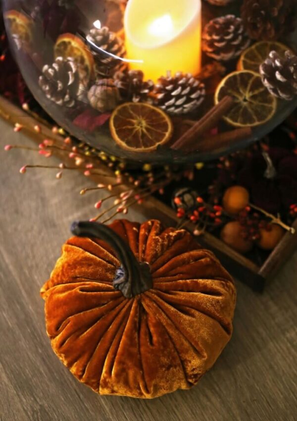 A plush velvet pumpkin sits on a wooden surface next to a glass bowl filled with a lit candle, dried orange slices, pinecones, and autumn decor.