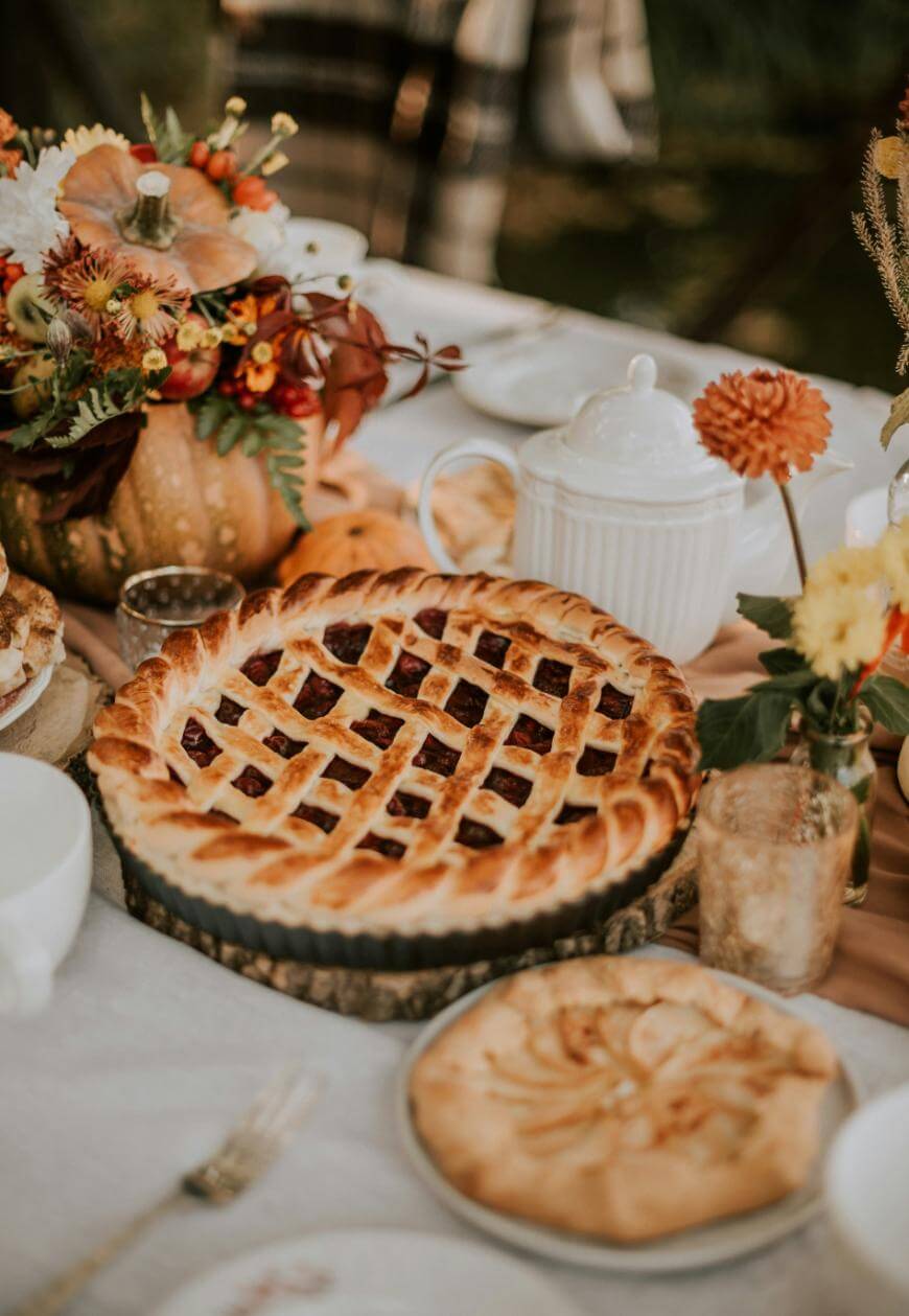 A table set for autumn with a lattice-topped pie, a smaller pie, a white teapot, glassware, and a pumpkin floral centerpiece decorated with fall flowers and leaves.