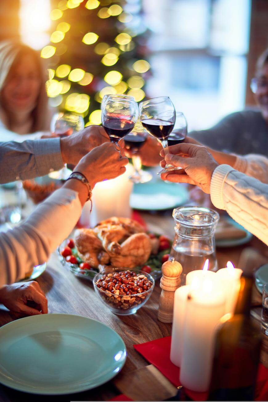 A group of people clink wine glasses over a festive dinner table decorated with candles, food, and a Christmas tree with lights in the background.