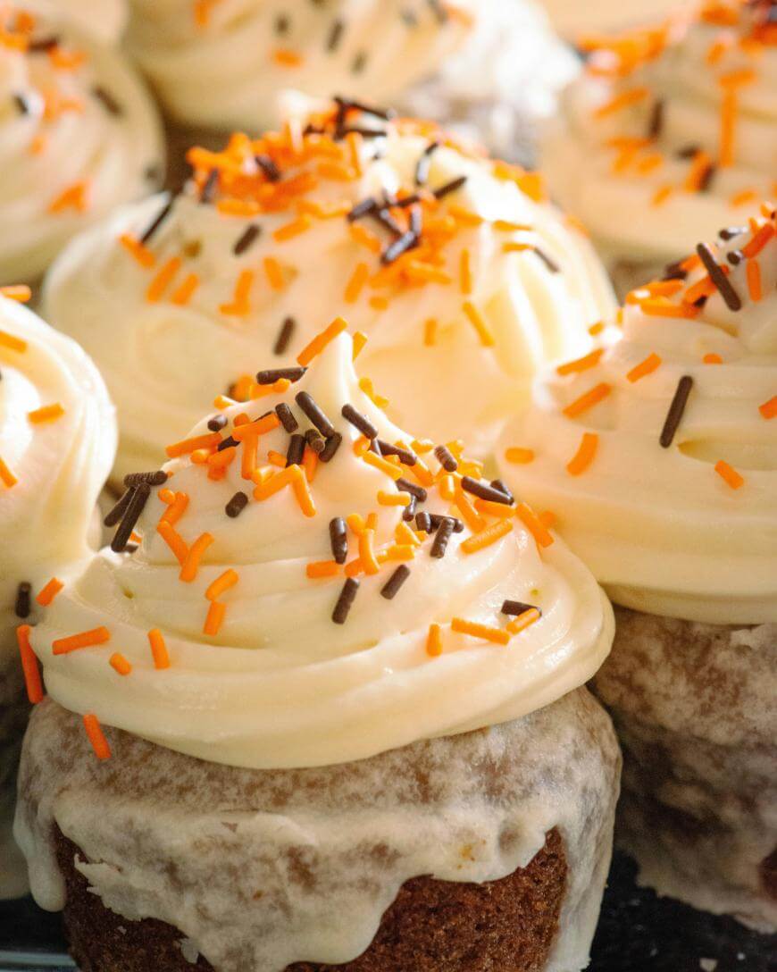 Close-up of cupcakes topped with swirled white frosting and orange and brown sprinkles. The cupcakes are arranged closely together, showcasing their festive decoration.