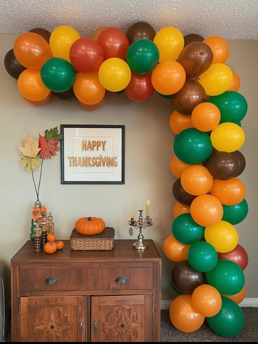 A fall-themed balloon arch in orange, yellow, green, and brown stands over a wooden cabinet decorated with pumpkins, leaves, and candles. A sign reading Happy Thanksgiving hangs on the wall behind.
