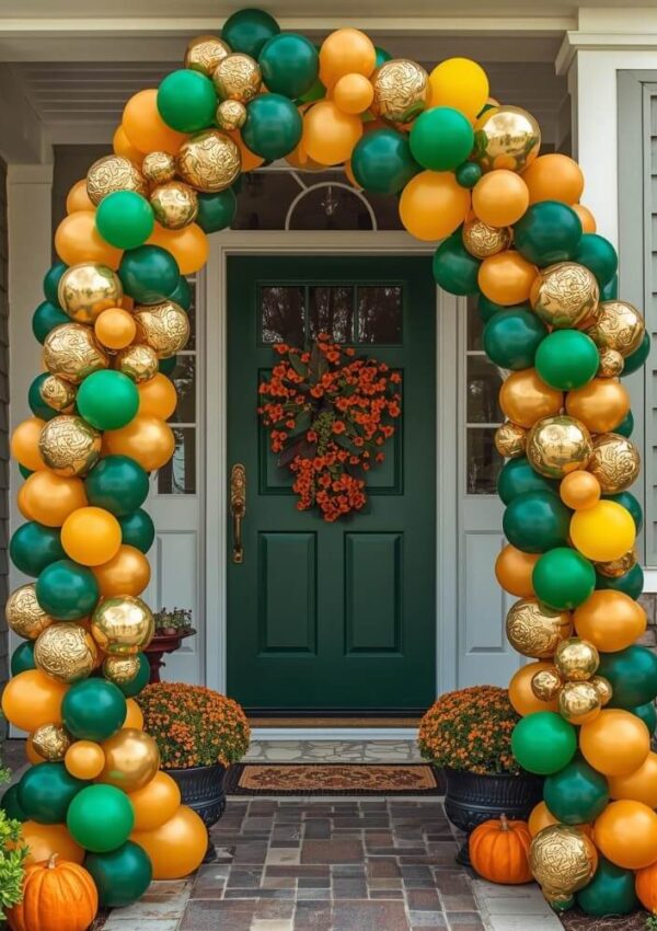 A green front door is framed by an archway of gold, orange, and green balloons. The porch is decorated with orange flowers, pumpkins, potted mums, and a wreath of orange berries on the door.