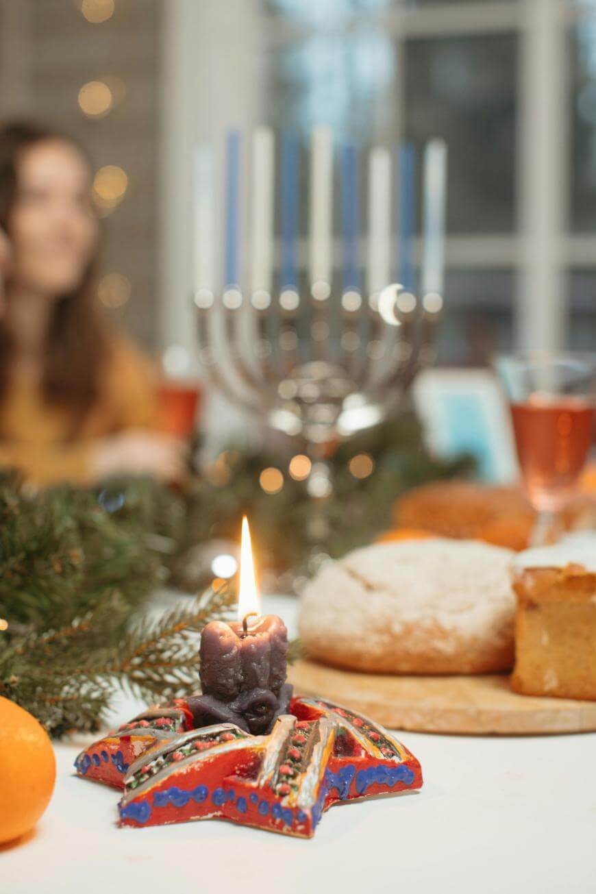 A lit candle in a decorated holder sits on a festive table with bread, drinks, and greenery. A menorah with blue and white candles is in the background, and a person is sitting out of focus.
