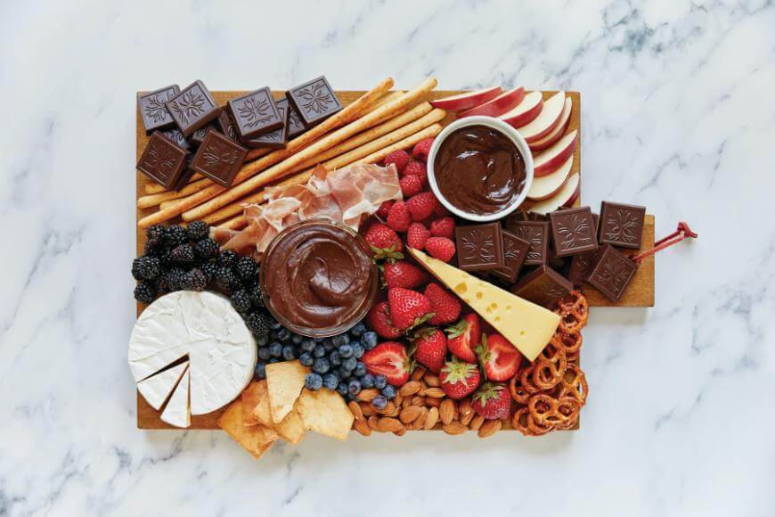 A wooden board topped with chocolate squares, breadsticks, sliced apples, strawberries, blackberries, blueberries, pretzels, crackers, almonds, cheese wedges, prosciutto, and two bowls of chocolate dip on a marble surface.