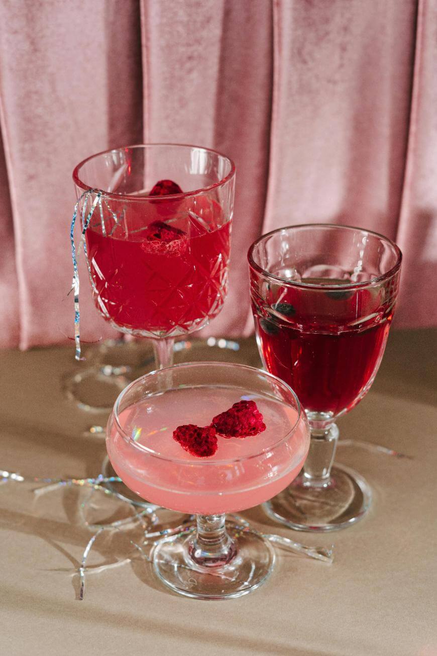 Three elegant glasses filled with pink and red drinks, each garnished with raspberries, are arranged on a table with metallic ribbon, set against a pink velvet backdrop.