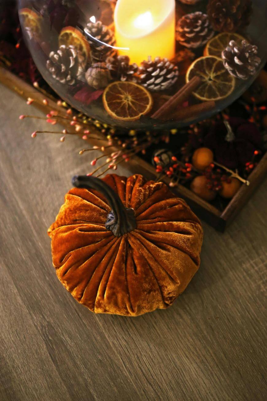 A plush velvet pumpkin sits on a wooden table next to a decorative tray with dried oranges, pinecones, berries, and a lit candle inside a glass bowl.
