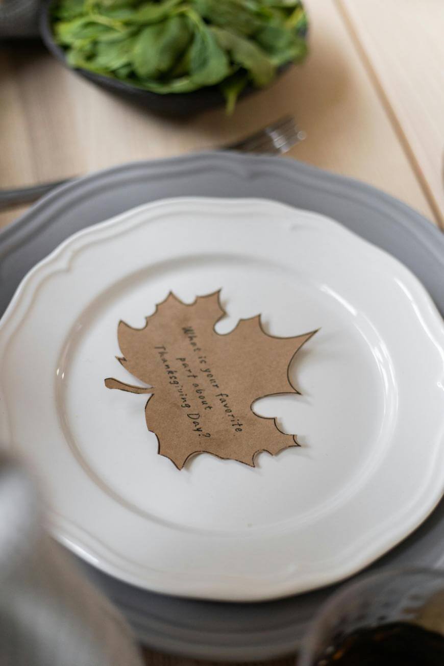 A white plate with a gray charger holds a paper cutout shaped like a maple leaf, printed with a Thanksgiving Day prompt. A fork and a bowl of greens sit nearby on a light wooden table.