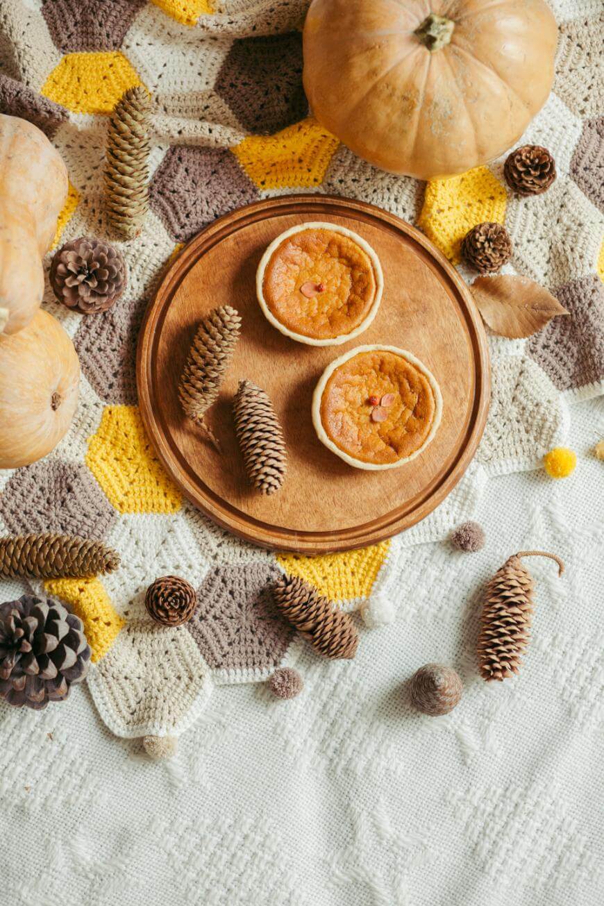 Two small pumpkin pies on a wooden plate with pinecones, pumpkins, and autumn decor on a patterned crochet tablecloth in cream, yellow, and brown tones.