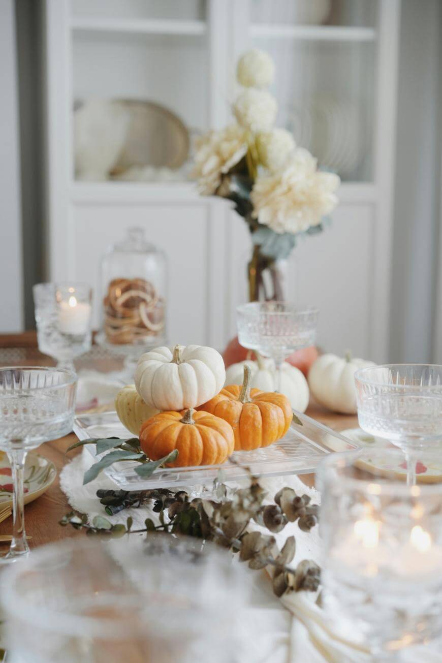 A close-up of a dining table decorated with small white and orange pumpkins, crystal glasses, candles, and a vase of white flowers, creating an elegant autumn or Thanksgiving centerpiece.