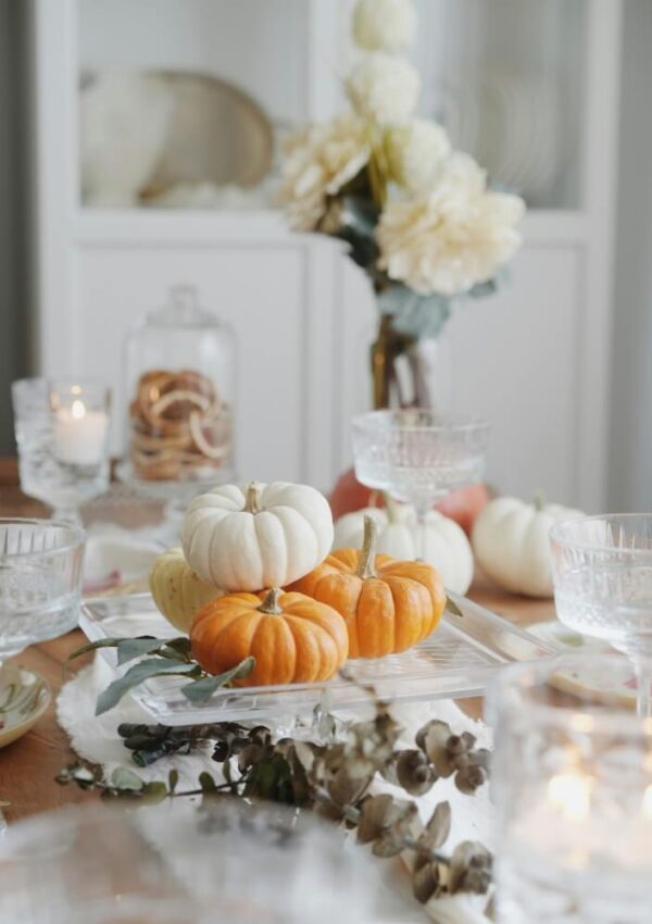 A fall-themed table setting with small white and orange pumpkins, crystal glassware, candles, dried foliage, and a vase of cream-colored flowers in the background. The scene feels elegant and festive.