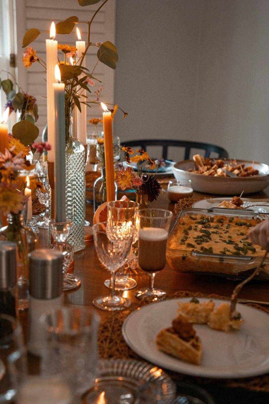 A warmly lit dining table set for a meal, featuring lit candles, floral arrangements, glassware, and dishes of food including a casserole and pie, creating a cozy, inviting atmosphere.