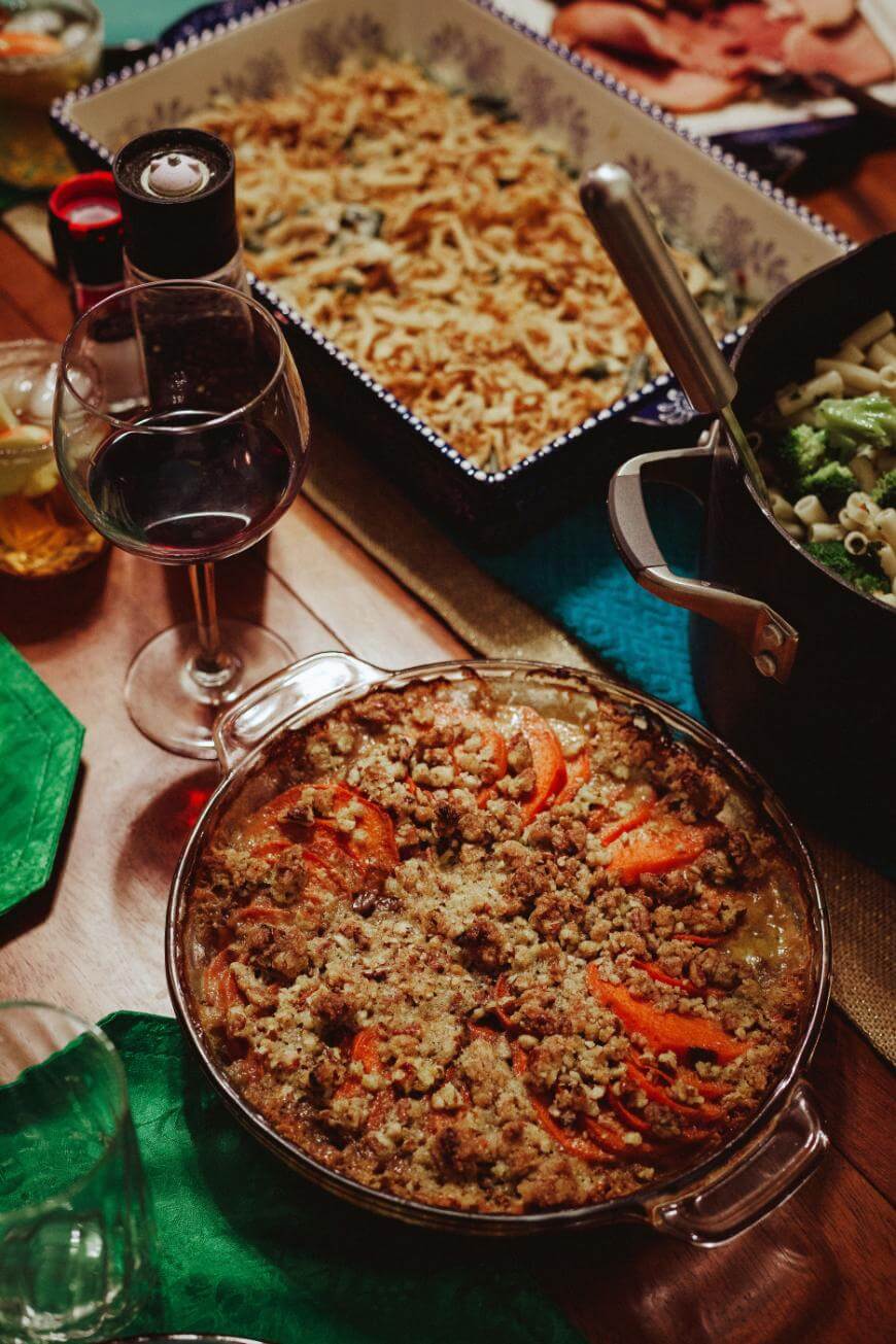 A glass of red wine sits next to a round dish of baked casserole topped with sliced tomatoes and crumbs, with a pot of pasta and a rectangular tray of green bean casserole nearby on a wooden table.