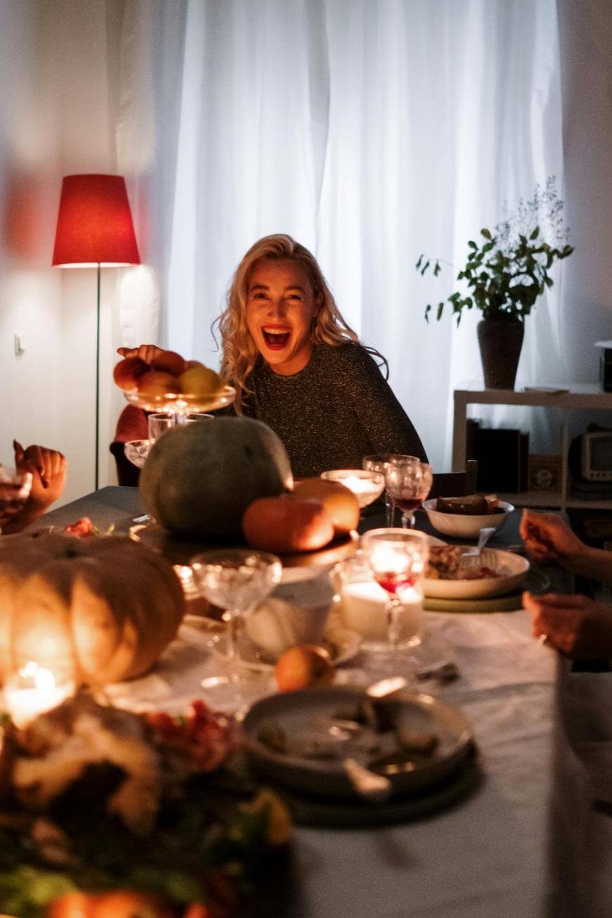 A woman with long blonde hair smiles and laughs at a candlelit dinner table, surrounded by food, pumpkins, and people. Warm lighting and a red lamp create a cozy atmosphere.