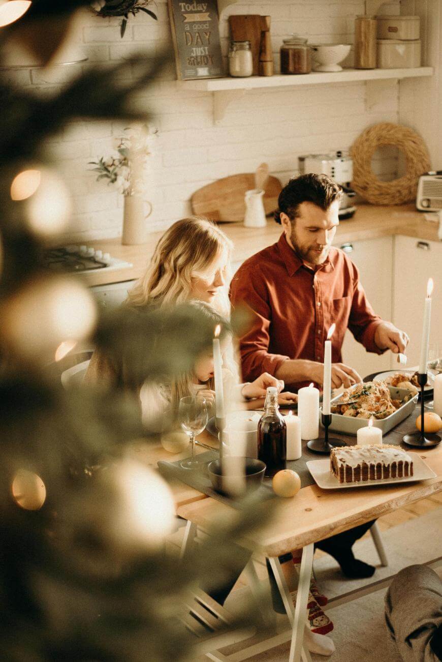 A man and woman sit at a wooden table enjoying a meal together in a cozy, warmly lit kitchen. A decorated Christmas tree is blurred in the foreground, and candles and festive food are on the table.