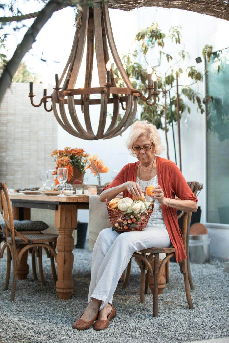 An elderly woman sits on a wooden chair outdoors, holding a basket of gourds. She is dressed casually with a red cardigan and glasses. Behind her is a wooden table set for a meal and a large rustic chandelier overhead.