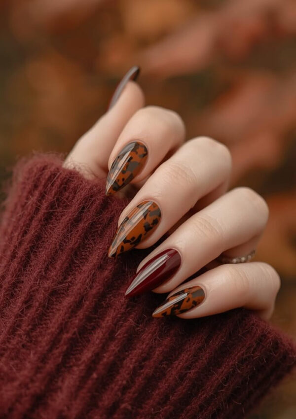 A hand with long, pointed nails features a fall-inspired manicure with maroon polish and tortoiseshell patterns. The person is wearing a cozy, ribbed burgundy sweater. The background is softly blurred in warm brown tones.