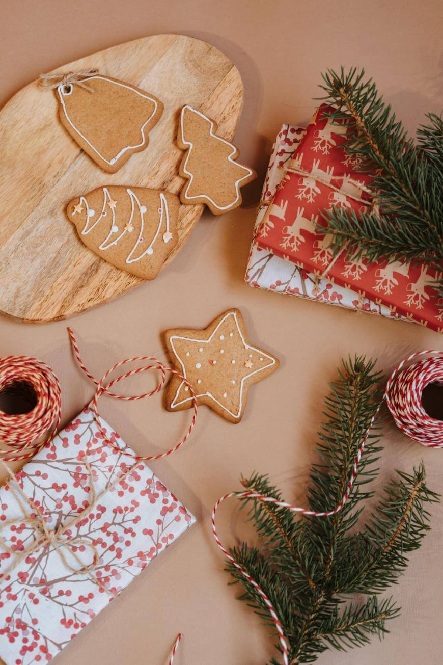 A festive scene with iced gingerbread cookies shaped like a bell, tree, and star on a wooden board, wrapped gifts, pine branches, and red-and-white twine on a brown surface.