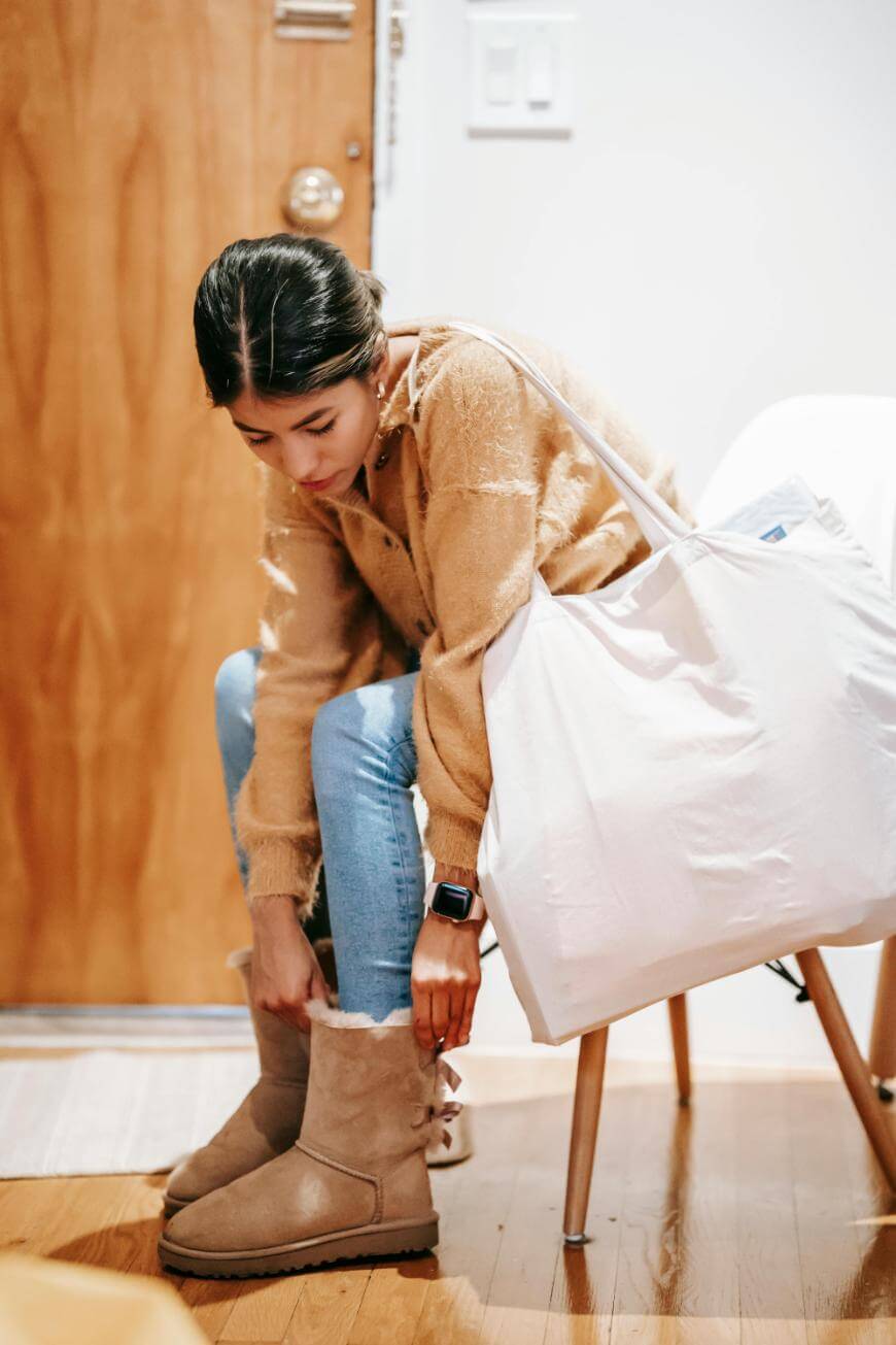 A woman sits on a chair, wearing a tan sweater and jeans, pulling on tan shearling boots. She has a large white tote bag over her shoulder and appears to be getting ready to go outside.