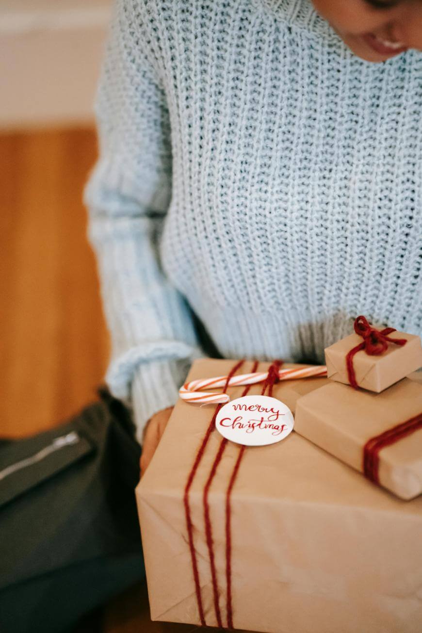 A person in a light blue knit sweater holds brown paper-wrapped gifts tied with red string. A candy cane and a tag reading Merry Christmas sit on top of one present.