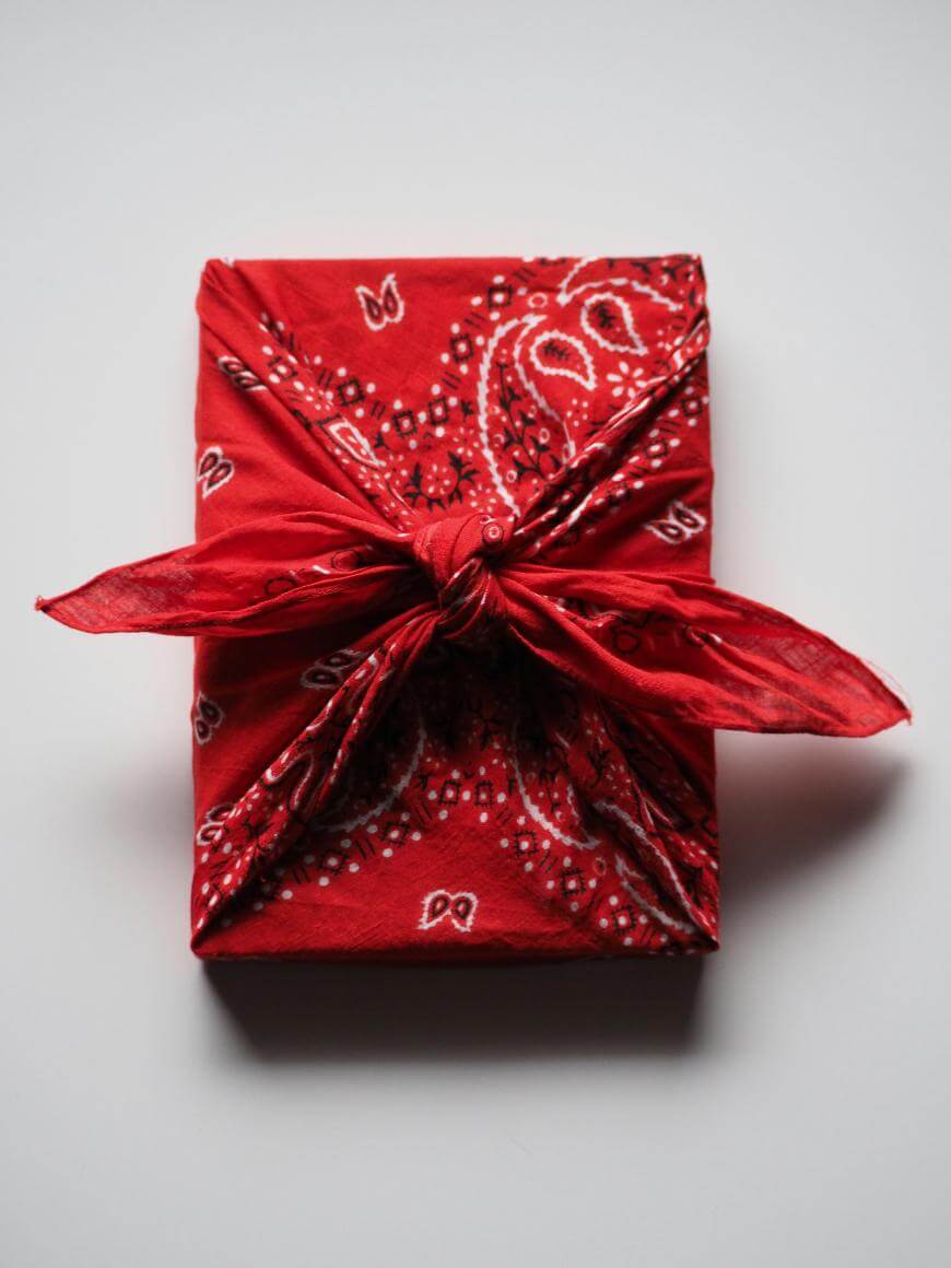 A rectangular gift wrapped in a red bandana with a paisley pattern, tied in a neat knot on top, set against a plain white background.