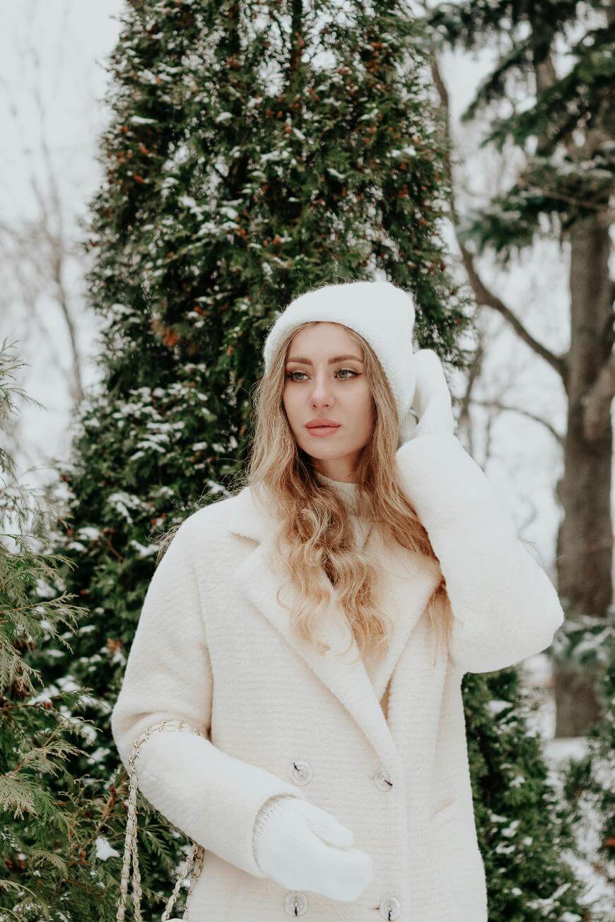 A woman with long blonde hair wearing a white coat, white knit hat, and white mittens stands outdoors in a snowy, green pine tree setting, looking thoughtfully into the distance.