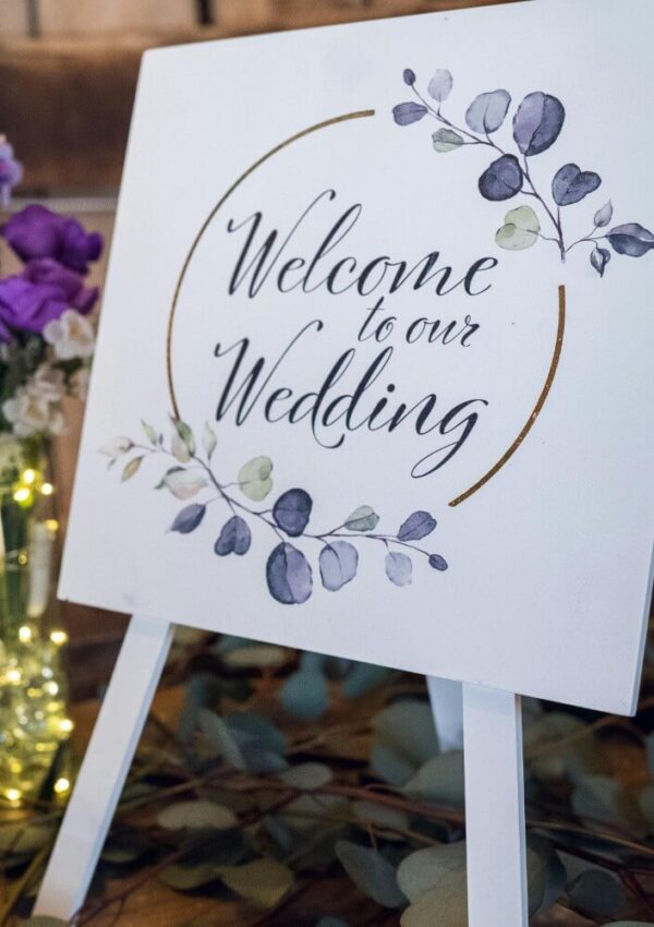 A white sign on an easel reads “Welcome to our Wedding” with watercolor-style leaves. In the background, there are purple flowers in a vase and string lights.
