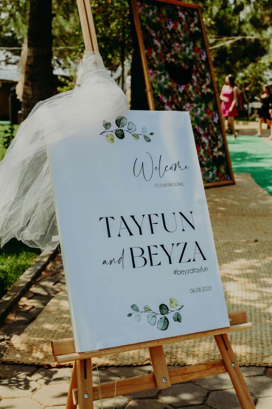 A white wedding welcome sign on an easel, decorated with tulle and floral designs, reads: Welcome to our beginning. Tayfun and Beyza. #beyzatayfun. 06.06.2022. Outdoors, with trees and people in the background.