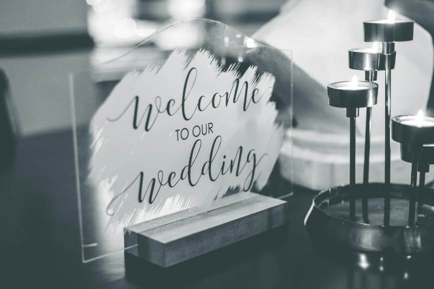A clear acrylic sign with the words Welcome to our wedding in elegant script stands on a wooden base next to lit candles. The image is black and white.