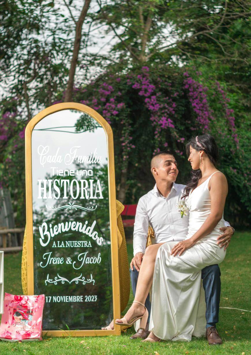 A couple dressed in formal attire sit together on a bench outdoors, smiling at each other. Next to them is a large decorative mirror with Spanish wedding text, surrounded by greenery and purple flowers.