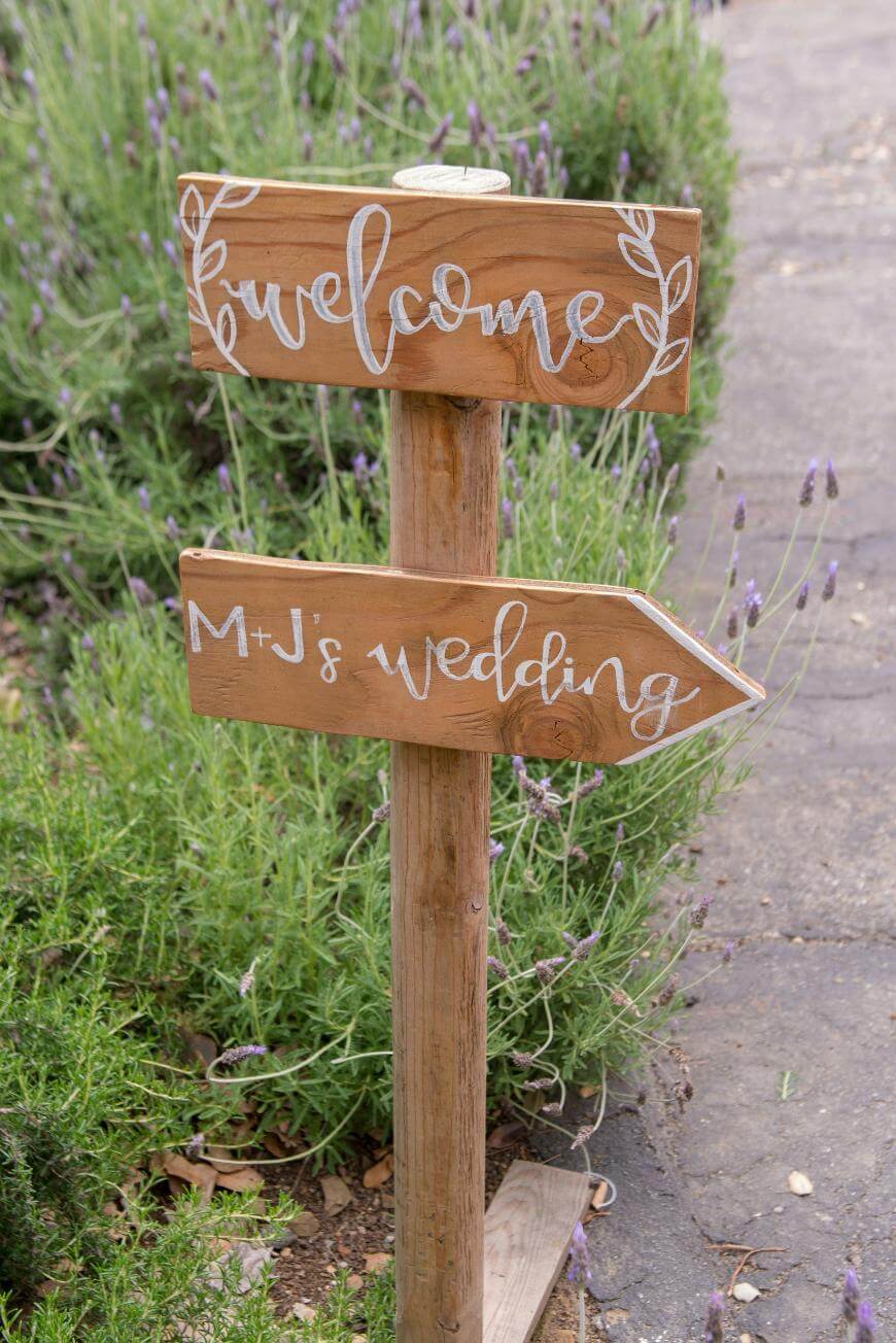 A wooden signpost with two signs, one reading “welcome” and the other pointing right with the words “M+J’s wedding,” stands in a garden with lavender plants and a stone path.
