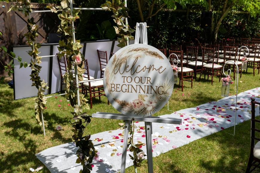 A decorative wedding sign on an easel reads Welcome to our beginning in a garden setting with rows of chairs, flower petals on a white aisle runner, and greenery surrounding the area.