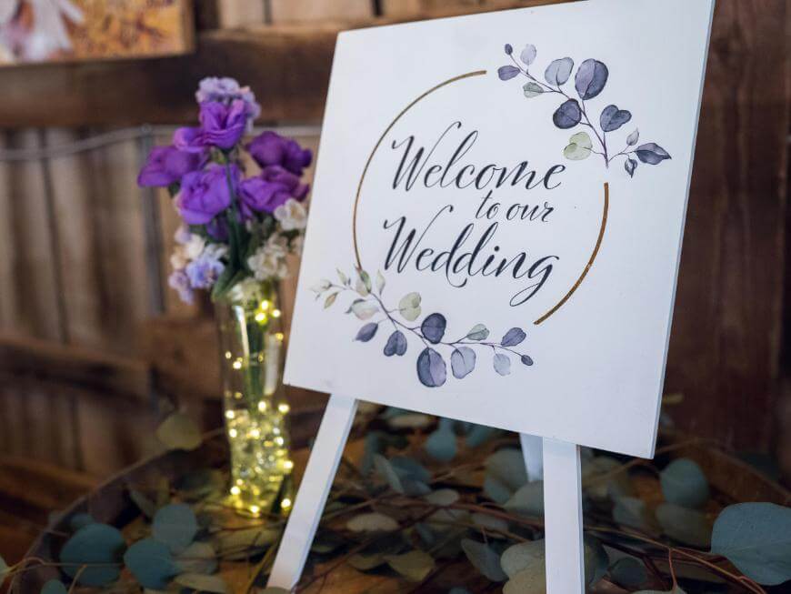 A white sign with floral decorations reads Welcome to our Wedding on an easel. Nearby, a glass vase with purple flowers and string lights sits on a rustic wooden table.