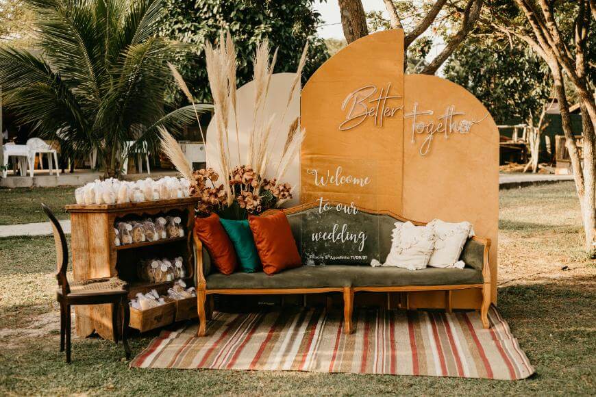 A wedding welcome area outdoors with a vintage green sofa, colorful pillows, pampas grass decor, a rug, a wooden side table with wedding favors, and a wooden backdrop sign reading Better together and Welcome to our wedding.