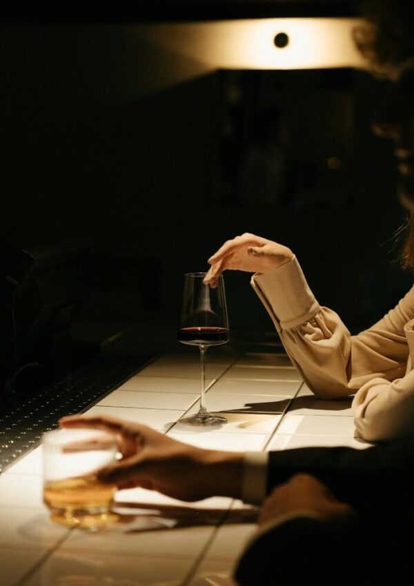 Two people sit at a dimly lit bar, each holding a drink—one with a glass of wine, the other with a glass of whiskey—resting their arms on a white tiled counter.