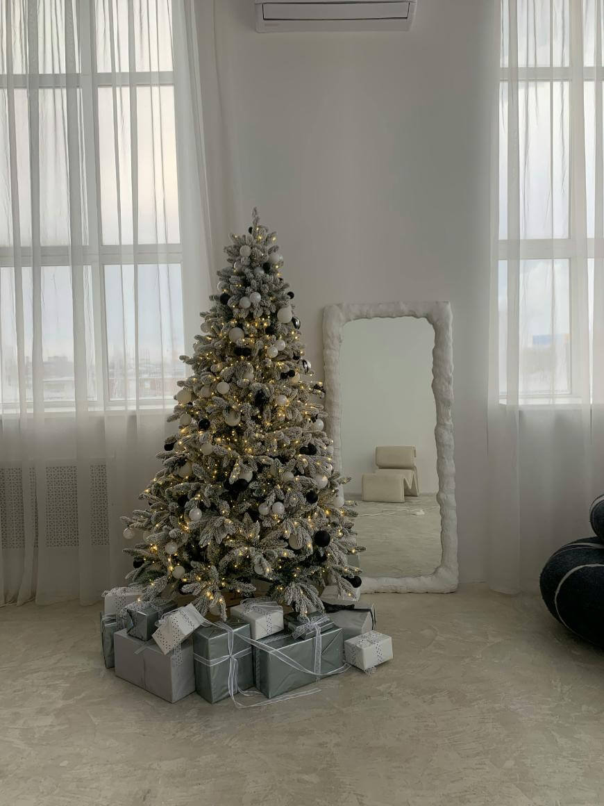A decorated Christmas tree with silver and gold ornaments stands in front of a large window, surrounded by silver-wrapped presents. A large mirror and a black chair are in the background in a bright, minimalist room.