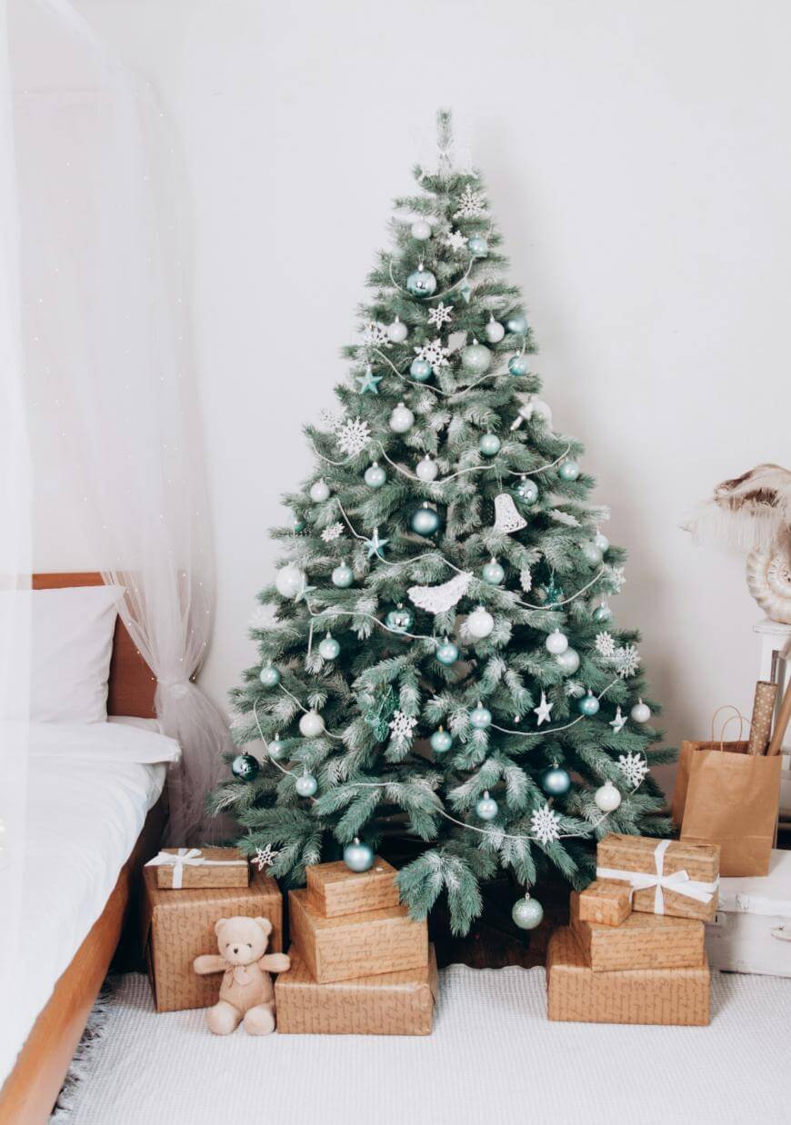 A decorated Christmas tree with white and silver ornaments stands beside a bed. Wrapped presents with white ribbons and a small teddy bear sit on the floor in front of the tree.