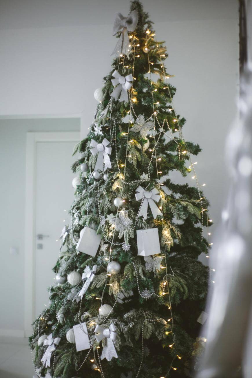 A Christmas tree decorated with white bows, silver ornaments, string lights, and white gift tags stands in a modern, minimalistic room with pale walls and a closed door in the background.