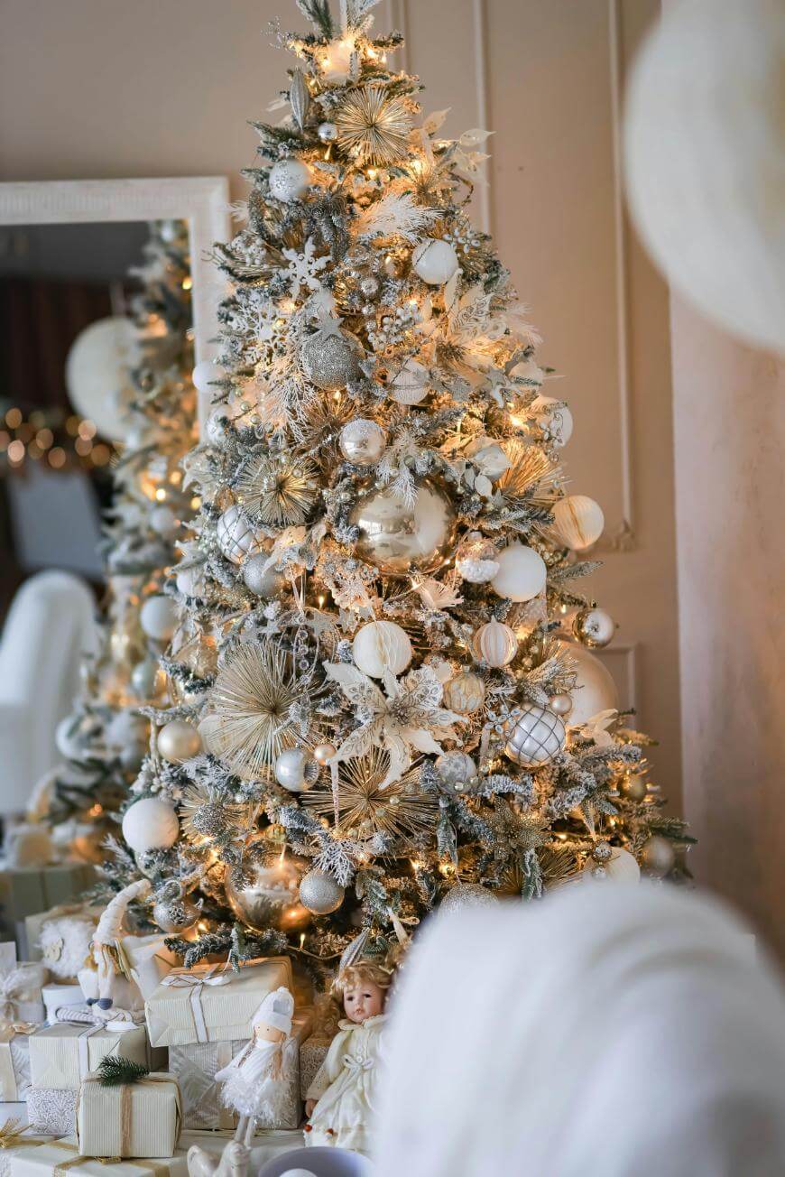 A Christmas tree decorated with white and gold ornaments, twinkling lights, and topped with snow-like accents. Wrapped presents and festive dolls are arranged underneath the tree in a cozy, elegant room.