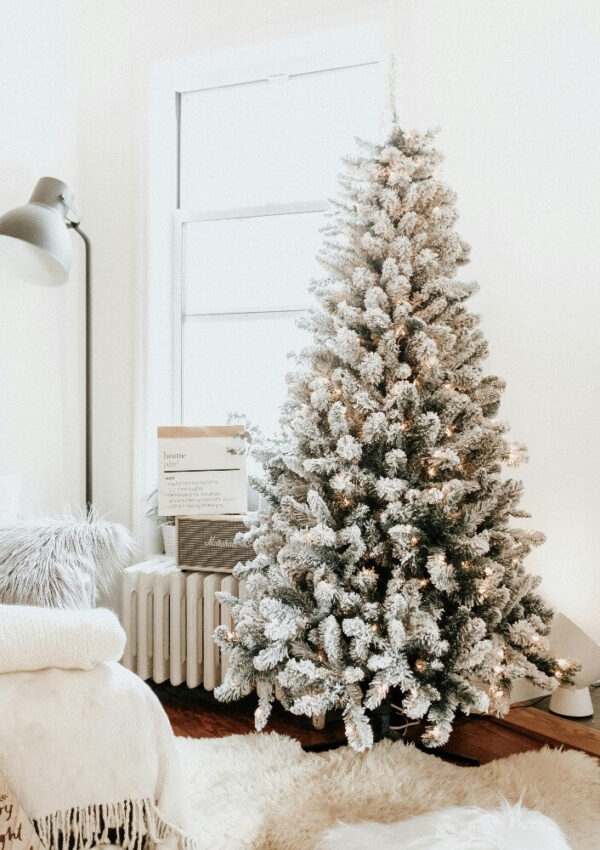 A snow-frosted Christmas tree with warm lights stands beside a window in a cozy, softly lit living room, decorated with fur throws, a fluffy rug, and a floor lamp. Wrapped gifts sit under the tree.