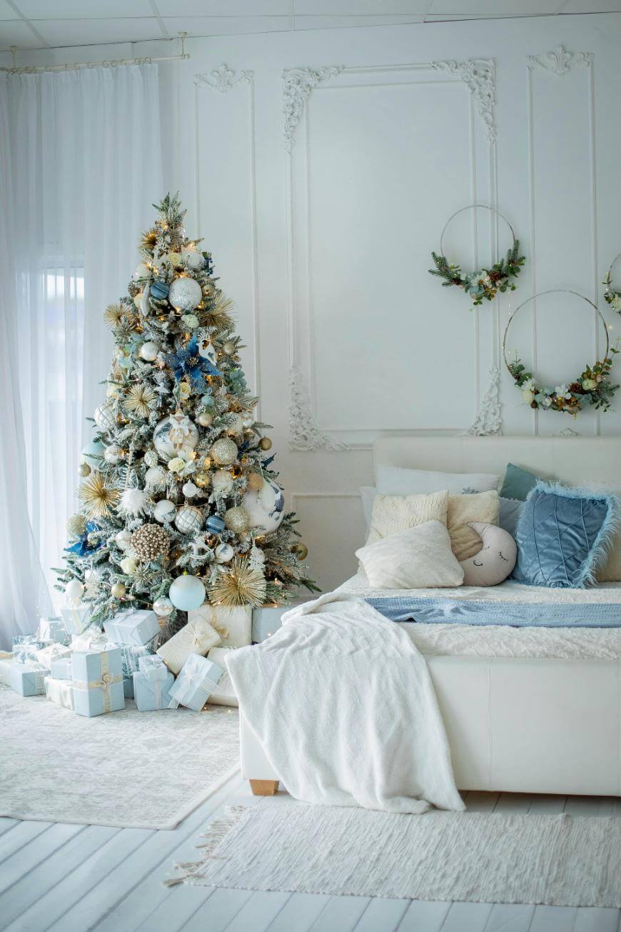 A beautifully decorated Christmas tree with blue and gold ornaments stands beside a white bed in a light, elegant room. Wrapped gifts are under the tree, and the bed has cozy pillows and blankets in white and blue tones.