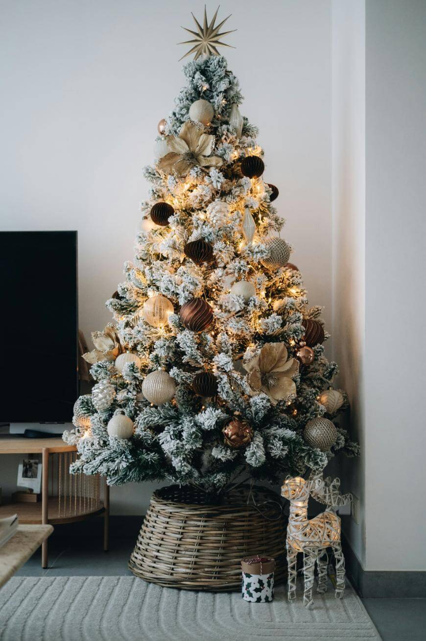 A decorated Christmas tree with white flocked branches, gold and brown ornaments, and warm lights stands in a wicker basket. A small decorative reindeer and a gift box sit at its base in a modern living room.