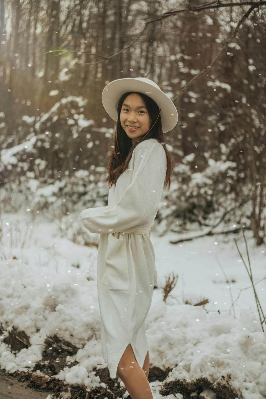 A young woman in a white dress and hat stands outdoors in a snowy forest, smiling at the camera as snow falls around her.
