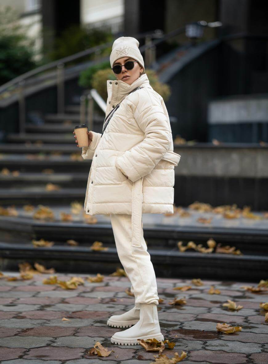 A person wearing a white puffer jacket, matching pants, boots, beanie, and sunglasses stands outdoors on a leaf-strewn walkway, holding a coffee cup and looking over their shoulder.