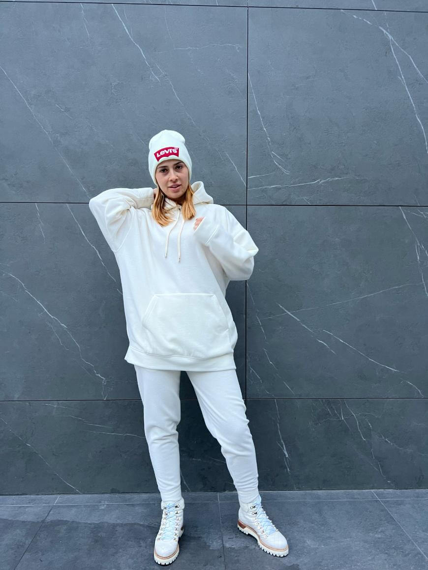 A woman poses against a gray marble wall, wearing a white beanie, oversized white hoodie, matching joggers, and white boots with a chunky sole. Her left hand is behind her head and her right arm rests at her side.