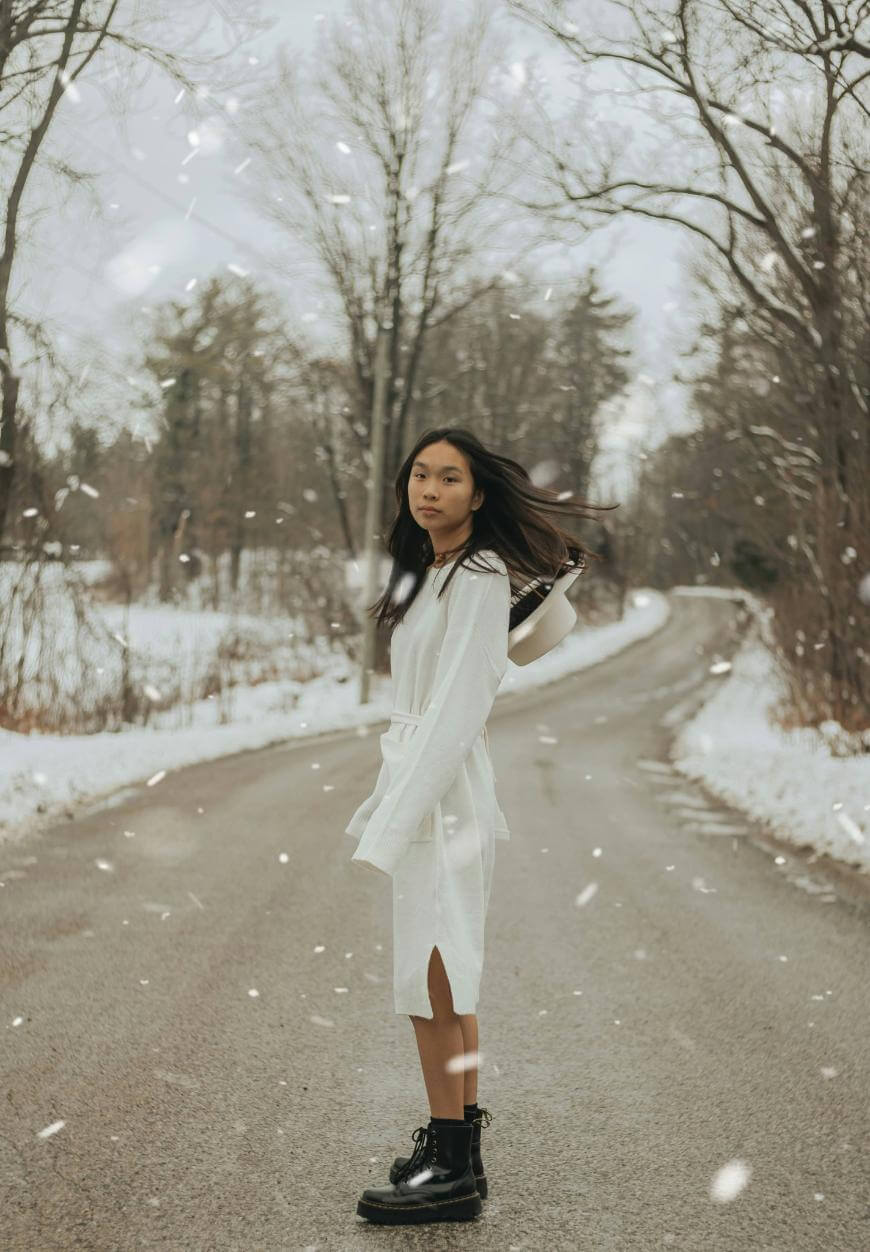 A young woman in a white dress and black boots stands on a snowy road lined with bare trees, looking at the camera as snow falls gently around her.